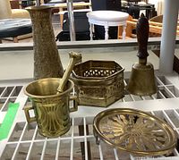 Full view of the brass decor items including vase, mortar and pestle, basket, bell, and trivet on shelf