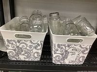 Two white plastic bins with decorative floral pattern holding multiple clear glass canning jars, some with metal lids.