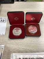Two silver dollar Canadian collector coins in individual display cases with red velvet interior, placed on a light wood surface along with a printed label.