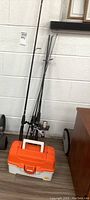 Three fishing rods standing upright leaning against a wall with the orange and white Plano tackle box in front of them.