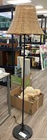 Full view of the metal floor lamp with a wicker shade in a retail setting, showing overall height and lamp design.