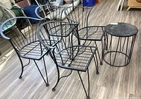 Set of four black metal patio chairs and one round metal side table displayed on wood-like floor, chairs and table show rust spots