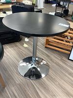 Full view of round black laminate top pedestal table with chrome base on hardwood floor, showing table top and surrounding area.