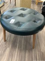 Front diagonal view showing black faux leather button tufted round stool with four natural wood legs.