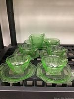 Photo showing four green uranium glass teacups resting on matching saucers and the sugar bowl and creamer behind them on a black surface.