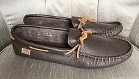 Side angled view of dark brown leather moccasins with tan leather laces tied in bows. Shows sole dimensions area and stitching details.