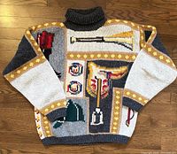 Front view of the sweater laid on floor showing colorful woven patches including motifs of a red boot, hunting horn, green hat, eagle emblem, and more.