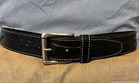 Close-up image of the black leather belt showing embossed details, white stitching, belt buckle, and leather loop with a stud.