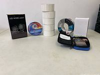 Overview of lot showing LED work light box, digital motor in case, multiple rolls of tape including white, black, and blue tapes.