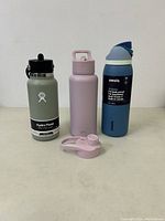 Three water bottles displayed together: Hydro Flask sage green, S'well Slim light purple with extra lid, Owala blue with straw.