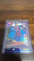 Holographic Panini Elite Lionel Messi football trading card showing Messi in FC Barcelona kit, card in protective sleeve, on wooden surface.