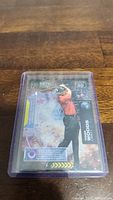 Photo of a Tiger Woods golf trading card from the Upper Deck Metal Universe series, showing the card inside a protective plastic sleeve on a wooden surface.