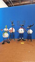 Four Christmas figurines on a brown surface against a blue background: three reindeer and one snowman, all with hollow bodies featuring star cutouts and cavities at the back for votive candles.