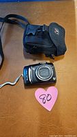 Canon PowerShot SX120 camera with wrist strap and black carrying case on brown surface with pink heart label '80'