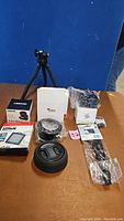 Photo showing Carson 4.5x 30mm LED sensor magnifier unopened box, Veal Tree camera lens with lens cap, small box, wrist strap in plastic wrap, and a tripod.