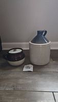 Two vintage ceramic crocks side by side on floor against wall, one lidded jar and one jug.