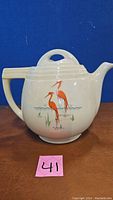 Front side of cream-colored ceramic coffee pot featuring painted design of two red-orange cranes standing in water with grass