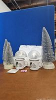 Full view of 3 silver bottlebrush trees, 2 silver brushed hemispherical light spheres, and 2 white ceramic Christmas mugs with festive prints, arranged on a wooden surface.