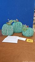 Front view of three teal wax pumpkins of varying sizes against a blue backdrop, showing detailed swirling patterns and leaf accents.