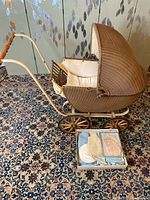 Side view of wicker baby carriage showing metal frame, wooden handle, wheels, and box of knit infant wear.