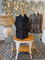 Three fur garments on mannequins: blonde mink stole on left, black and grey faux fur vest center, 1950s grey goat hair jacket right.