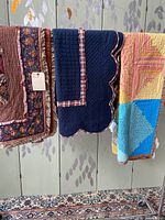 Three quilted coverlets hung side by side showing different colors and trim styles: floral and plaid, navy with plaid trim and scalloped edges, multicoloured log cabin pattern.