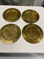 Four round brass charger plates arranged on a white tablecloth, showing surface oxidation and tarnish