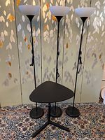 Three black metal torchiere floor lamps with white plastic shades, 69 inches tall, and one black adjustable computer table with triangular top and three-legged base on patterned rug.