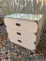 Off-white 3-drawer nightstand dresser with glass top and nickel plated handles against leafy wallpaper background on patterned carpet.
