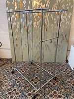 Photo showing the entire adjustable height double-sided metal rolling rack with caster wheels placed on a patterned carpet in front of a decorative screen.