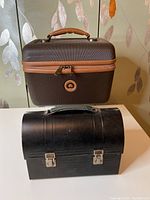 Photo showing black metal lunch box and brown Delsey traincase side by side