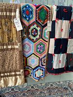 Three quilts and afghan hanging side by side. Left quilt in brown floral with satin band patchwork, middle colourful crochet hexagonal pattern afghan, right quilt with dark blue squares and decorative ties.