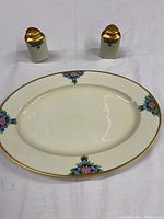 Oval serving dish with floral decoration and gold trim, photographed with salt and pepper shakers on white fabric background.