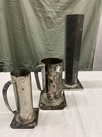 Three tin candle molds displayed on a white surface against a cloth background, two cylindrical with handles and one tall hexagonal mold.
