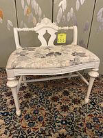 Front view of the white wooden vanity bench showing painted carved backrest and leaf patterned cushioned seat on a decorative leg structure.