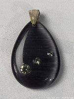 Close-up image showing smooth dark teardrop stone with three embedded clear faceted stones and attached metal bail.