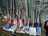 Wide view of 20 yard and garden tools standing against a weathered green wooden wall outdoors showing various shovels, forks, rakes, hoes, and axes
