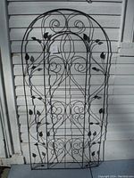Front view of the black metal garden trellis with scroll and leaf detail leaning against a white paneled wall.