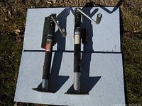 Top view of two black and silver heavy duty trailer jacks with crank handles placed side by side on a board outdoors.