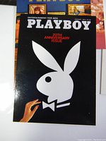 Close up of a Playboy magazine cover card showing the iconic white bunny logo on a black background with '20th Anniversary Issue' text.