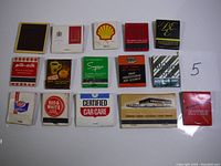 14 vintage advertising matchbooks laid out in rows on a white surface showing brand logos and text on covers.