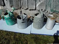 Four old galvanized watering cans and planters sitting on a blue surface outside by a weathered wooden wall, showing overall lot content.