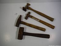 Four unique hammers on white surface with metal and wooden parts visible. The first hammer has a pronged head, the second a ball-peen head, the third a blacksmith hammer shape, and the fourth is a large sledgehammer-style.
