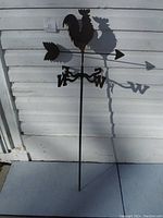 Full front view of the metal rooster weather vane showing its height and design against a white wall.