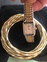 Close-up image showing gold-tone wristwatch with rectangular face and two-tone metal band resting on set of three twisted gold-tone bangle bracelets. Black textured background visible.