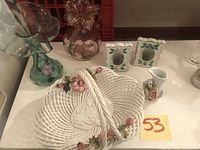 Front view of ceramic basket, pink and blue blown glass vases, porcelain floral frames and bud vase on shelf.