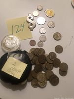 Collection of various coins arranged on a surface with black pouch labeled '2 Drachma Greece' and a silver dollar sign money clip.