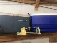 Front and side view of two large coolers and metal garden sprinkler placed on a wooden shelf.