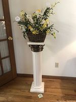 Full view of the white Corinthian style ceramic pedestal holding a wicker basket with artificial flowers, placed on wooden floor near a glass door and electrical socket in background