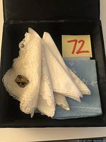 Top view of the box containing three white handkerchiefs with lace and embroidery and one light blue embroidered cloth, alongside an Empire style brooch